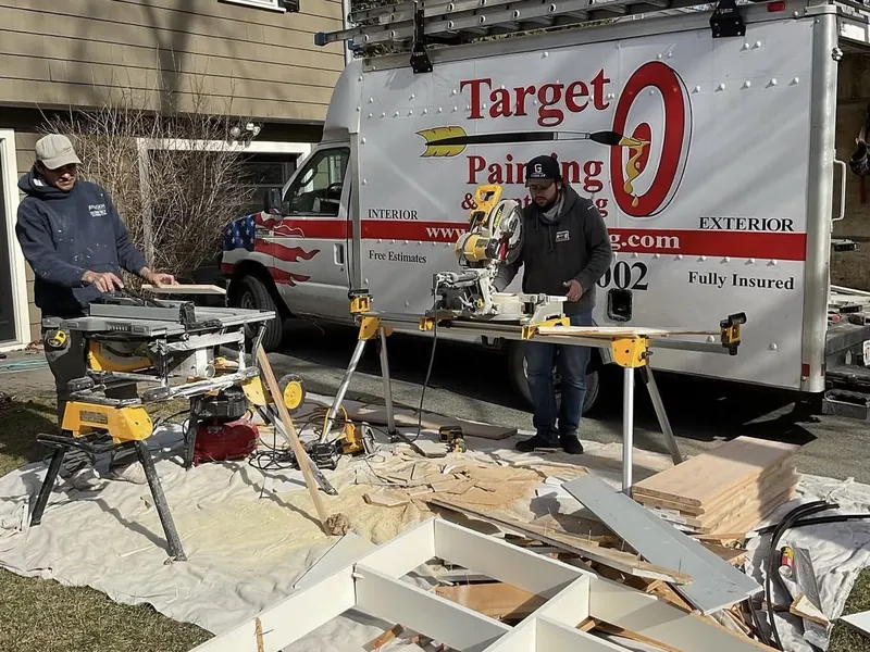 Two Target Painting carpenters cutting new trim on saws beside the company truck at a home