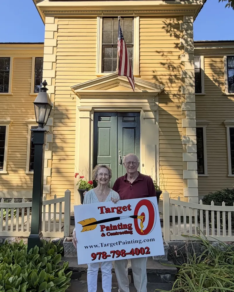 Smiling couple holding a Target Painting yard sign in front of their freshly painted yellow colonial