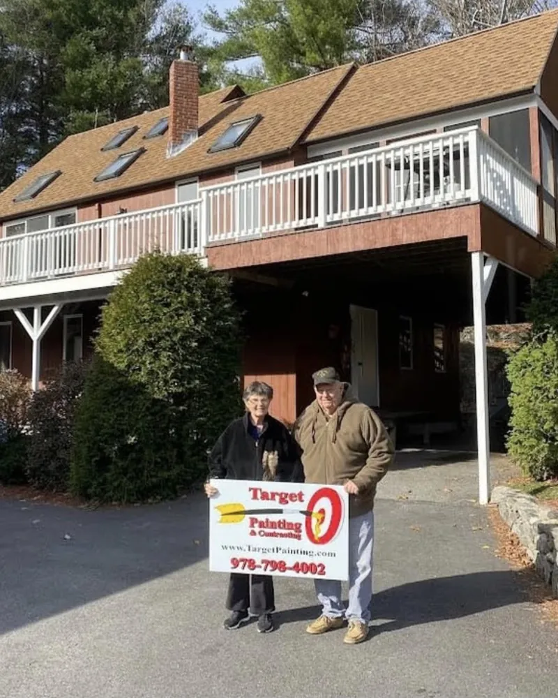 Two homeowners holding a Target Painting yard sign in the driveway of their painted home with a big wraparound deck